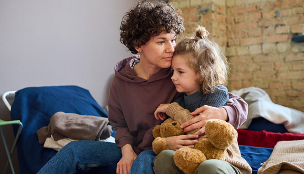 A woman holds a young child while both sit on a shelter bed